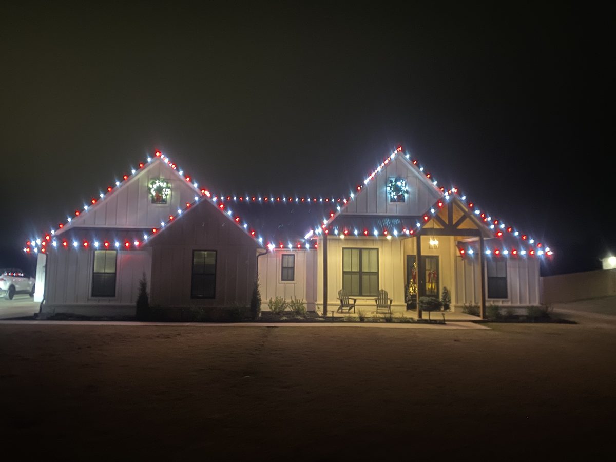 Farmhouse outlined in red and white Christmas lights with lit wreaths in the gables